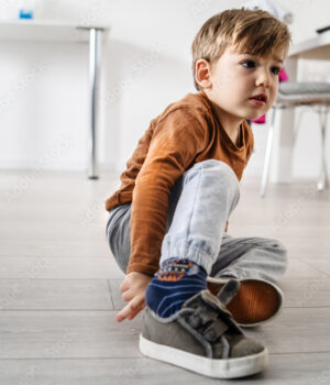 Full length view of small cute caucasian boy little child kid sitting on the wooden or laminated vinyl floor at home putting on or taking off the shoes in day alone front view